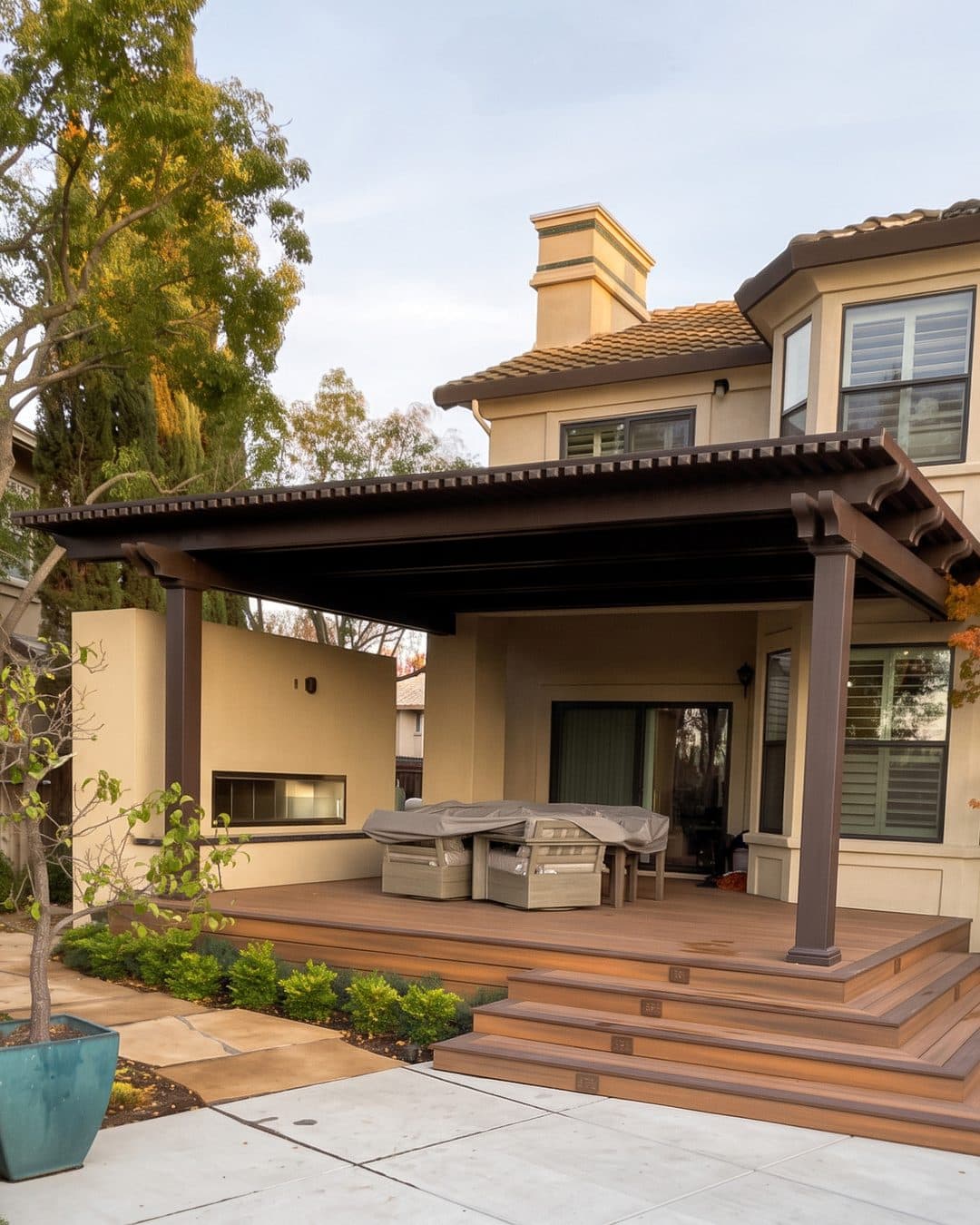 Bronze patio cover with rafter-tail trim over a raised composite deck and wide steps, beside a two-story home and a red Japanese maple