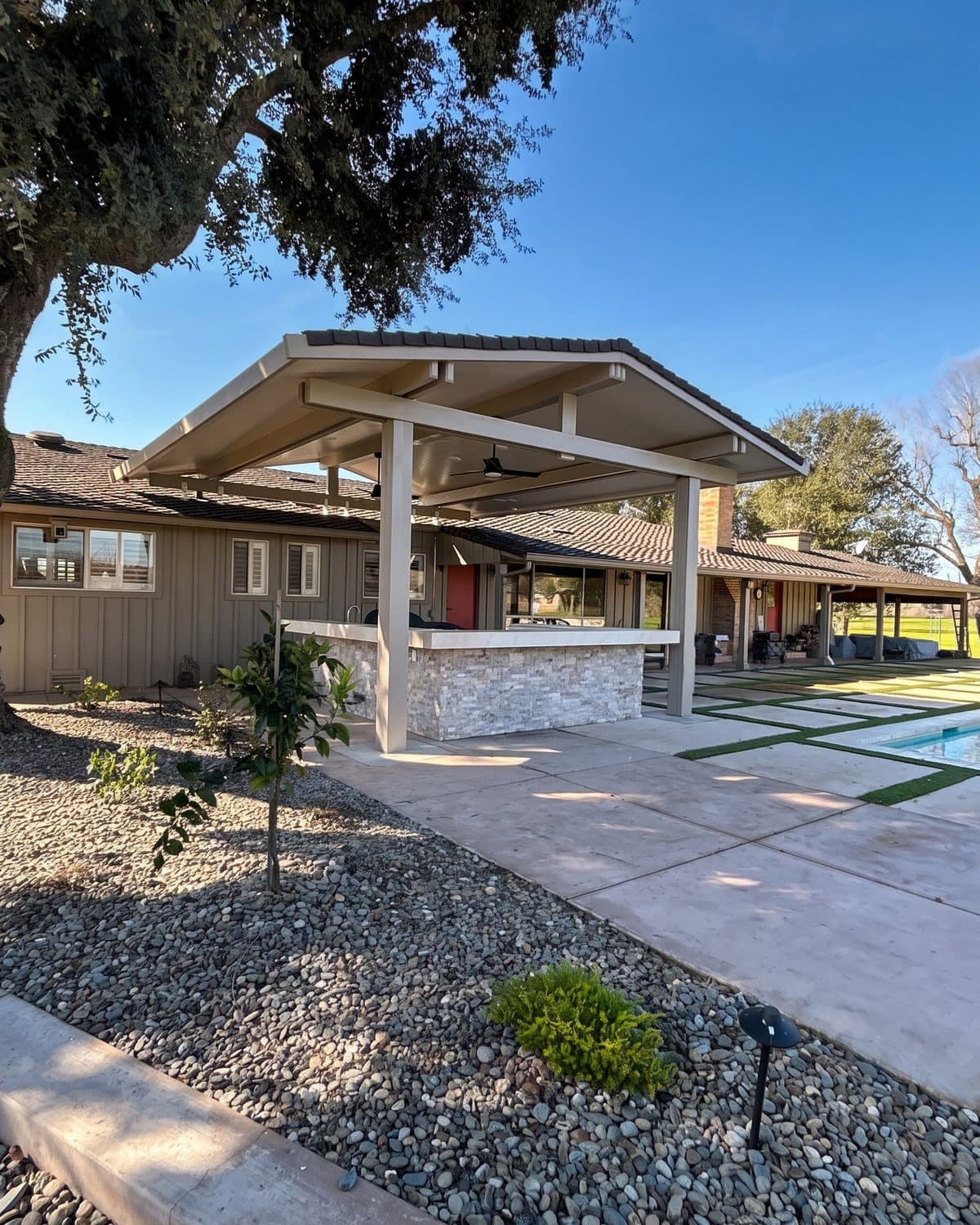 Freestanding white gabled patio cover over a stacked-stone outdoor kitchen beside the pool, under a spreading oak