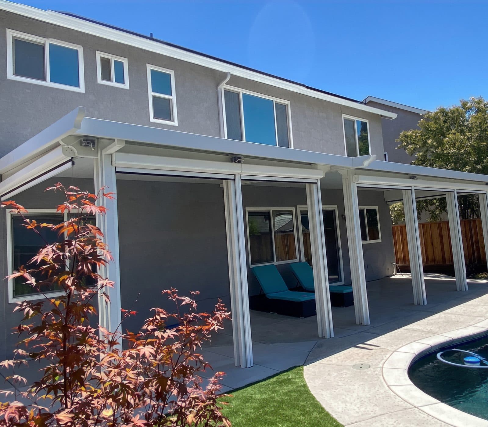 Long white patio cover on slim columns shading lounge chairs along a curved pool, with a red Japanese maple in front