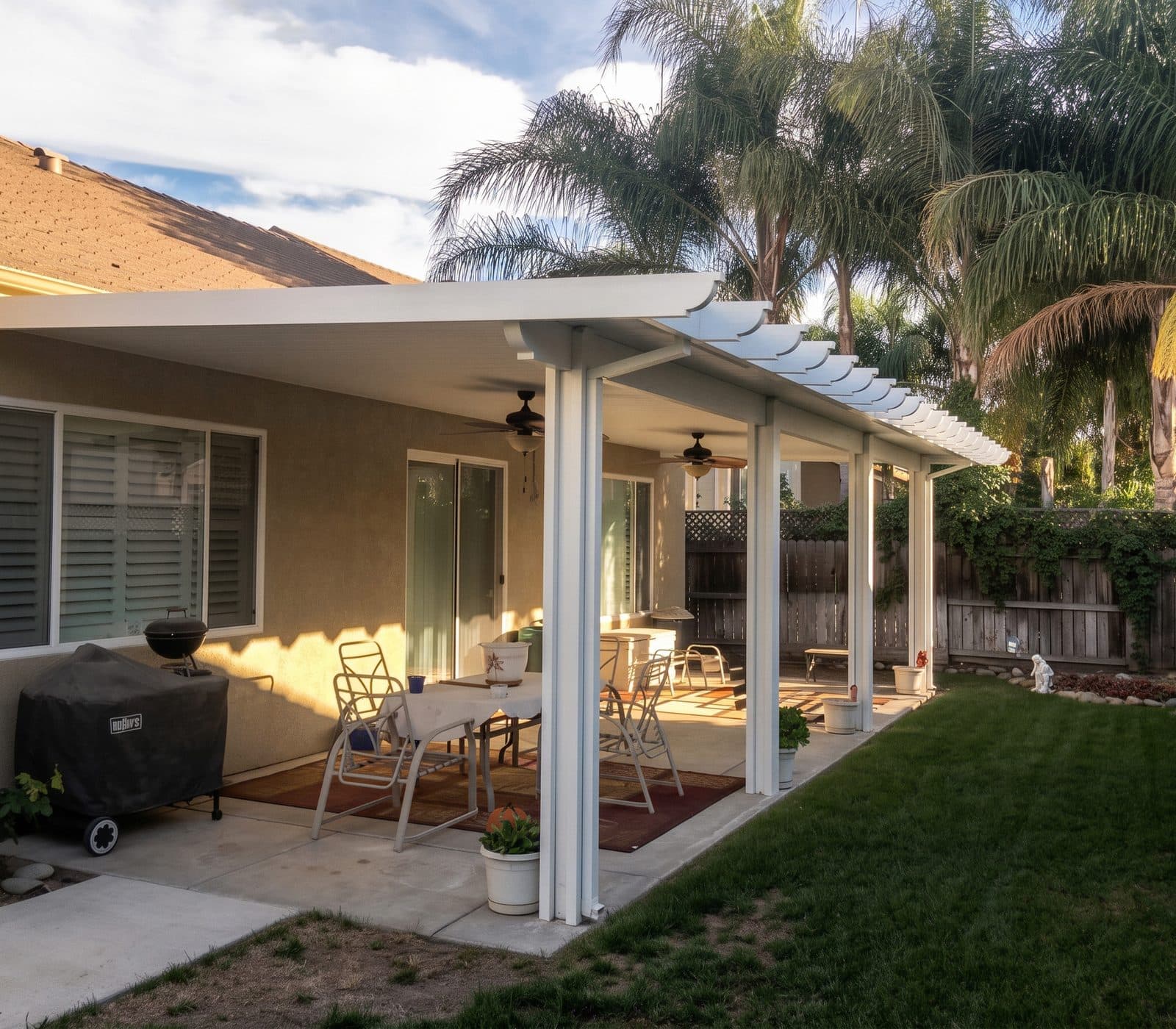 White insulated patio cover with lattice ends shading a backyard dining area under tall palms