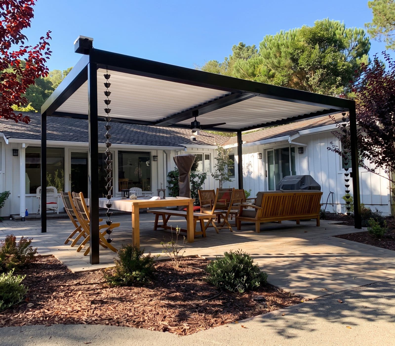 Black louvered pergola with copper rain chains over an outdoor dining table and lounge chairs behind a white farmhouse-style home