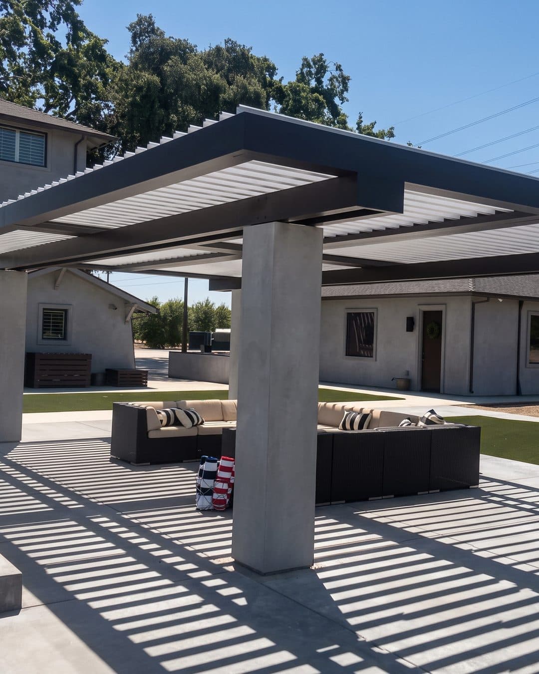Black-framed louvered pergola on concrete columns casting striped shade across an outdoor sectional and patio